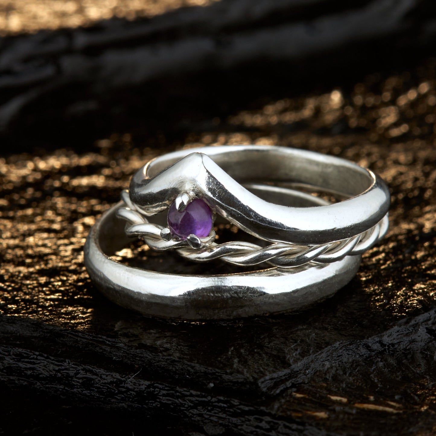 Handgefertigtes Ehering-Set aus 925 Silber auf Leder. 3-teiliges Damen-Set bestehend aus einem 3mm Wishbone-Ring mit Amethyst und einem 2mm Kordelring. Daneben ein massiver 4mm Herrenring im Halbrund-Profil. Zeitloses Design mit lila Edelstein für die Hochzeit. SEO-Keywords: Wishbone Eheringe Silber, Amethyst Trauringe Set, handgefertigte Silberringe, 925 Sterlingsilber Schmuck, Kordelring Hochzeit. Sichtbare Details der hochwertigen Handarbeit und Steinbesetzung.