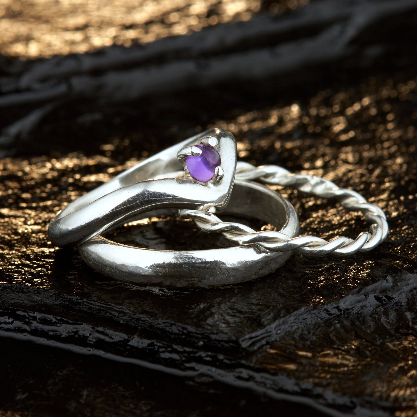 Handgefertigtes Ehering-Set aus 925 Silber auf Leder. 3-teiliges Damen-Set bestehend aus einem 3mm Wishbone-Ring mit Amethyst und einem 2mm Kordelring. Daneben ein massiver 4mm Herrenring im Halbrund-Profil. Zeitloses Design mit lila Edelstein für die Hochzeit. SEO-Keywords: Wishbone Eheringe Silber, Amethyst Trauringe Set, handgefertigte Silberringe, 925 Sterlingsilber Schmuck, Kordelring Hochzeit. Sichtbare Details der hochwertigen Handarbeit und Steinbesetzung.