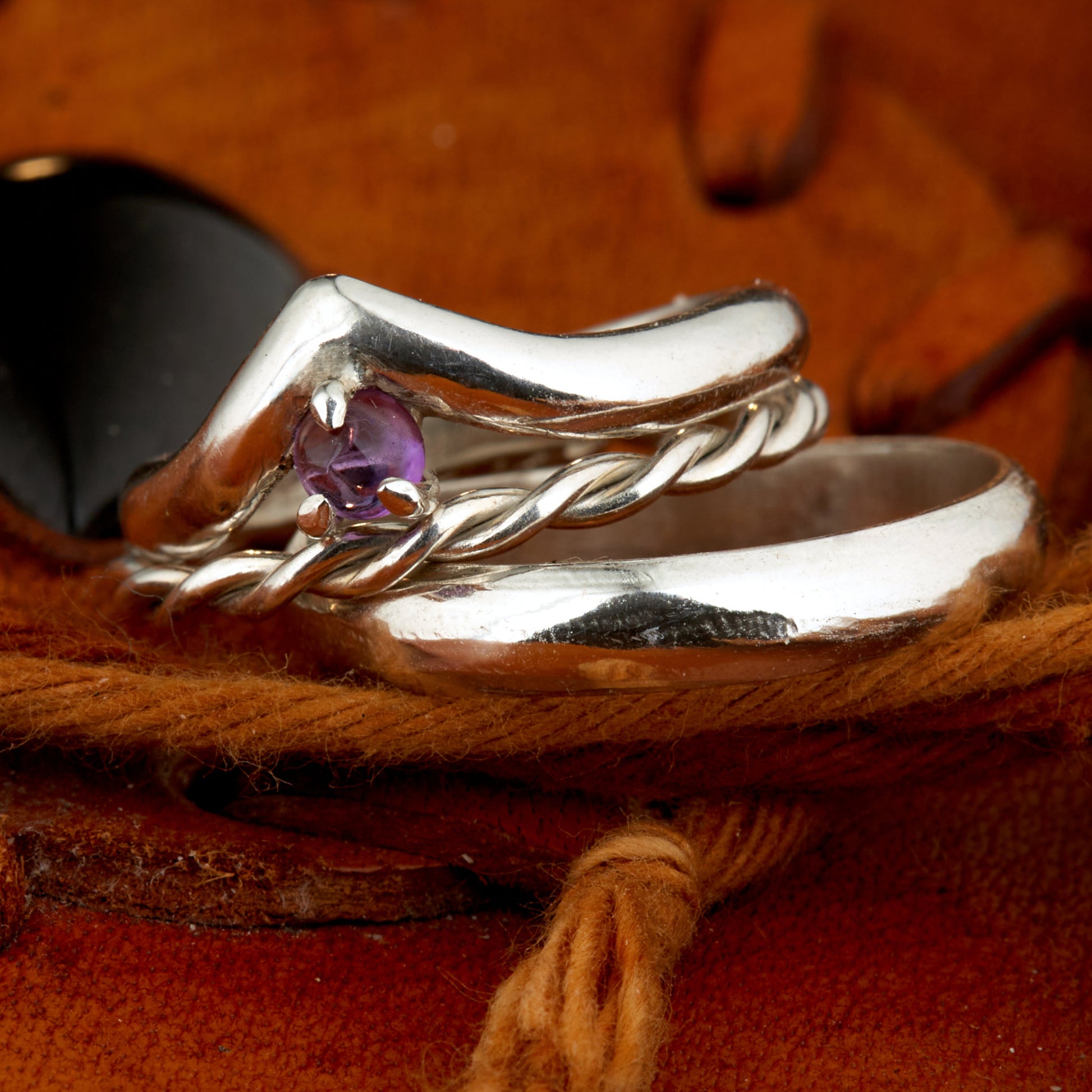 Handgefertigtes Ehering-Set aus 925 Silber auf Leder. 3-teiliges Damen-Set bestehend aus einem 3mm Wishbone-Ring mit Amethyst und einem 2mm Kordelring. Daneben ein massiver 4mm Herrenring im Halbrund-Profil. Zeitloses Design mit lila Edelstein für die Hochzeit. SEO-Keywords: Wishbone Eheringe Silber, Amethyst Trauringe Set, handgefertigte Silberringe, 925 Sterlingsilber Schmuck, Kordelring Hochzeit. Sichtbare Details der hochwertigen Handarbeit und Steinbesetzung.