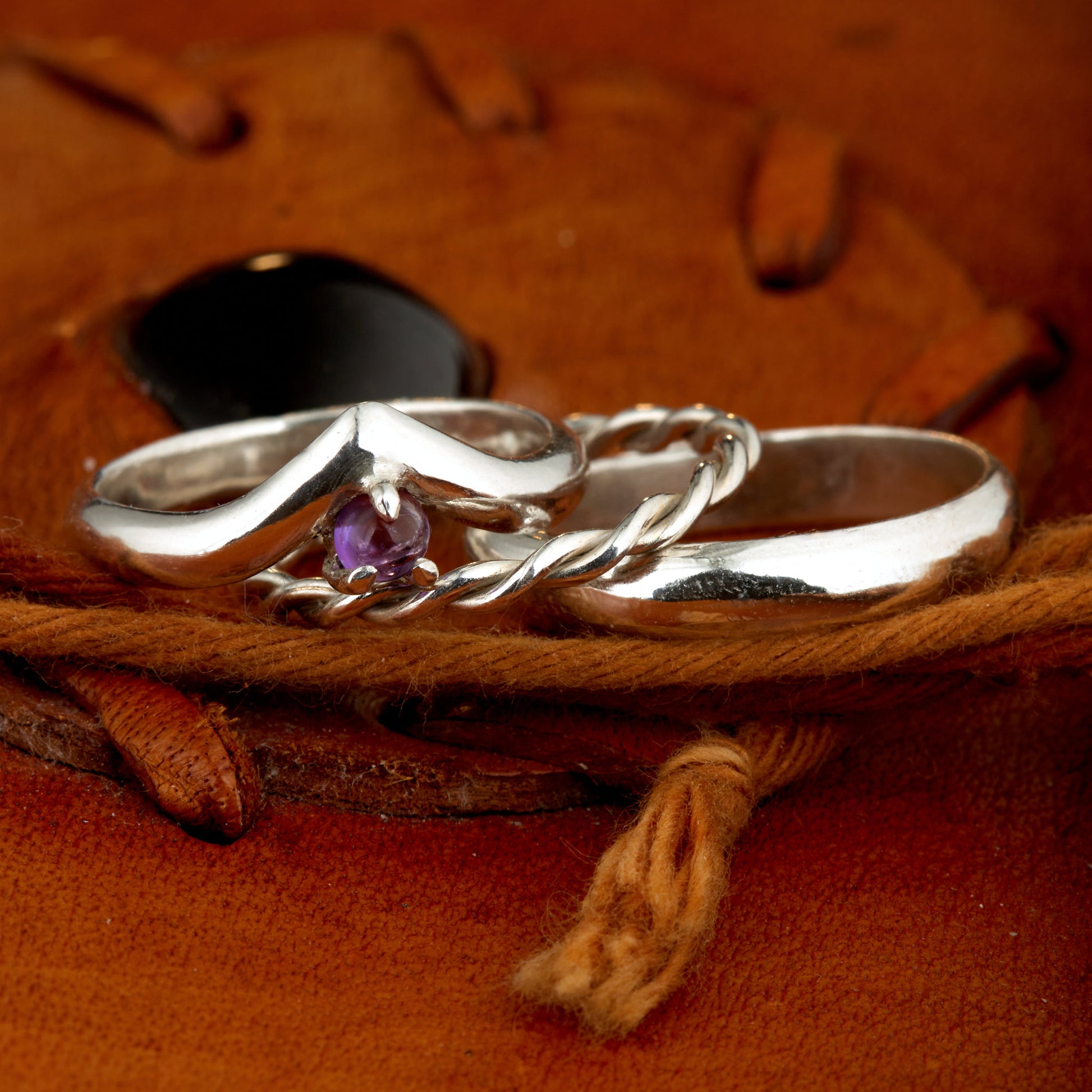 Handgefertigtes Ehering-Set aus 925 Silber auf Leder. 3-teiliges Damen-Set bestehend aus einem 3mm Wishbone-Ring mit Amethyst und einem 2mm Kordelring. Daneben ein massiver 4mm Herrenring im Halbrund-Profil. Zeitloses Design mit lila Edelstein für die Hochzeit. SEO-Keywords: Wishbone Eheringe Silber, Amethyst Trauringe Set, handgefertigte Silberringe, 925 Sterlingsilber Schmuck, Kordelring Hochzeit. Sichtbare Details der hochwertigen Handarbeit und Steinbesetzung.