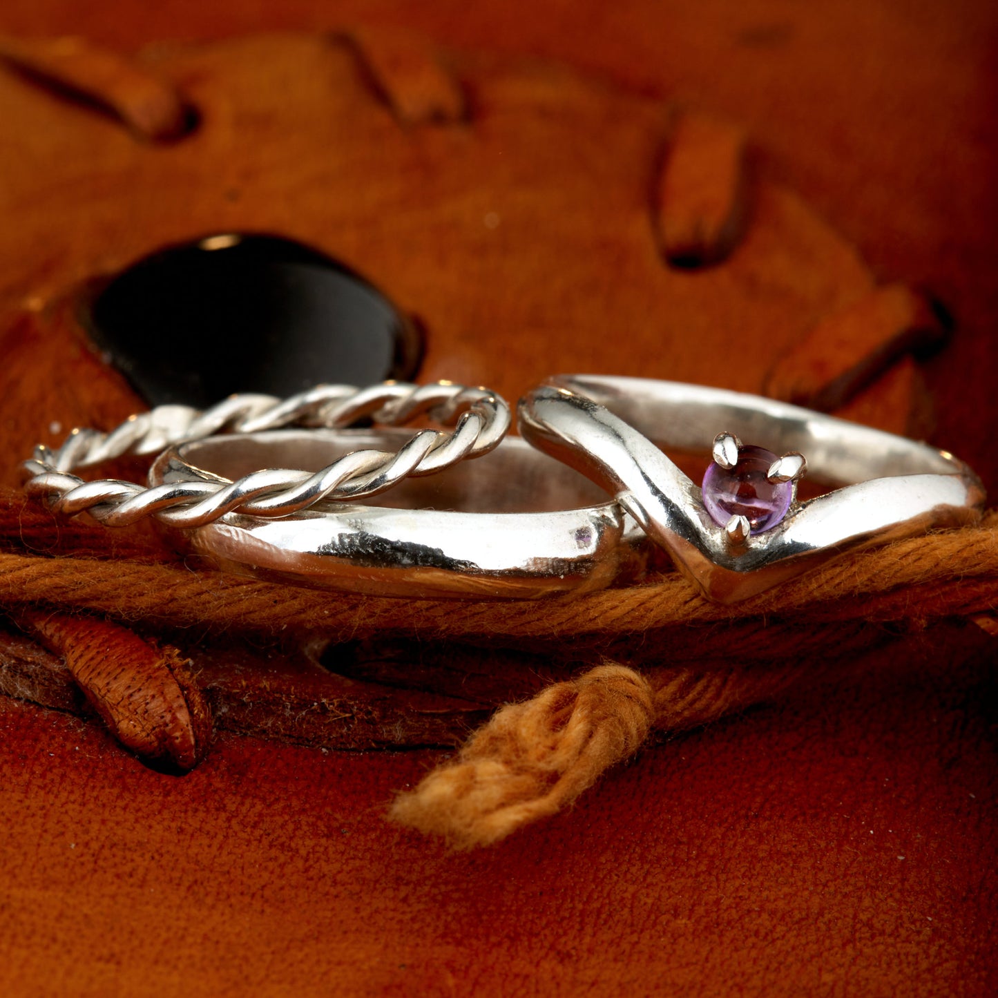 Handgefertigtes Ehering-Set aus 925 Silber auf Leder. 3-teiliges Damen-Set bestehend aus einem 3mm Wishbone-Ring mit Amethyst und einem 2mm Kordelring. Daneben ein massiver 4mm Herrenring im Halbrund-Profil. Zeitloses Design mit lila Edelstein für die Hochzeit. SEO-Keywords: Wishbone Eheringe Silber, Amethyst Trauringe Set, handgefertigte Silberringe, 925 Sterlingsilber Schmuck, Kordelring Hochzeit. Sichtbare Details der hochwertigen Handarbeit und Steinbesetzung.