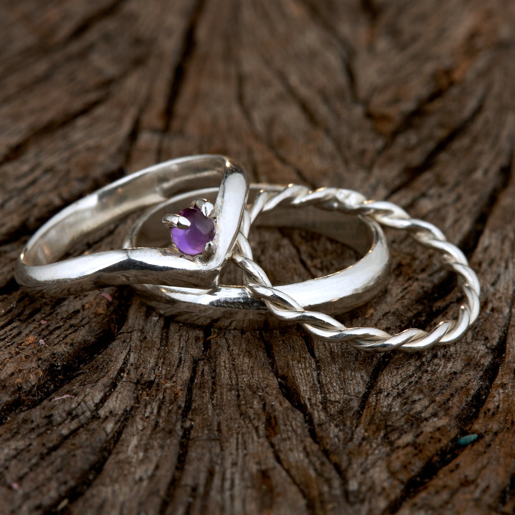 Handgefertigtes Ehering-Set aus 925 Silber auf Leder. 3-teiliges Damen-Set bestehend aus einem 3mm Wishbone-Ring mit Amethyst und einem 2mm Kordelring. Daneben ein massiver 4mm Herrenring im Halbrund-Profil. Zeitloses Design mit lila Edelstein für die Hochzeit. SEO-Keywords: Wishbone Eheringe Silber, Amethyst Trauringe Set, handgefertigte Silberringe, 925 Sterlingsilber Schmuck, Kordelring Hochzeit. Sichtbare Details der hochwertigen Handarbeit und Steinbesetzung.